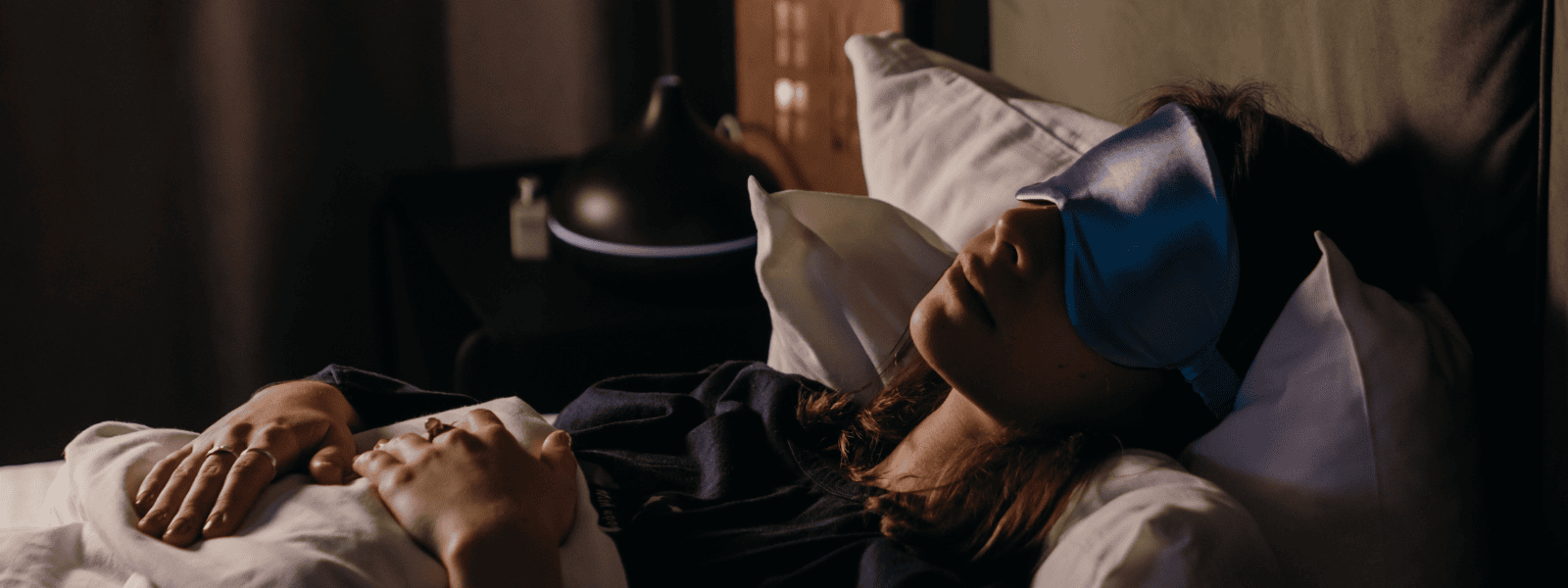 A woman wearing a sleep mask rests peacefully in bed, with soft lighting illuminating the room. 