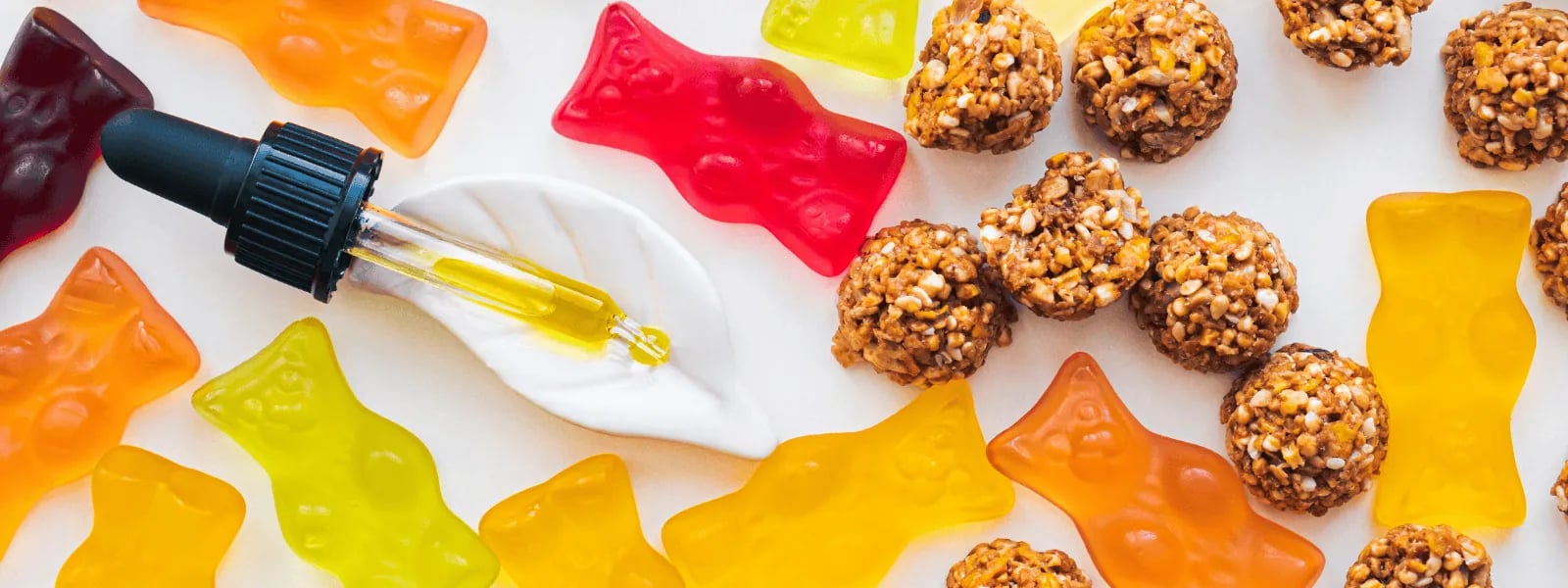 Colorful gummy candies and granola bites arranged around a dropper dispensing oil onto a dollop of cream on a white surface.